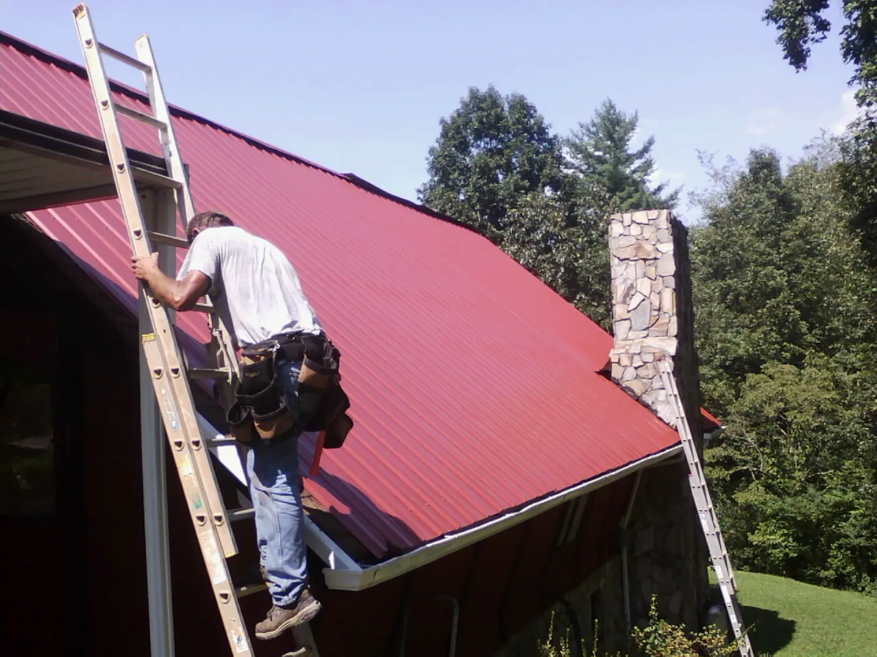 Roofer on a ladder beside a red metal roof and stone chimney — Soffit & Fascia Repair by Roofing Brothers in Pooler
